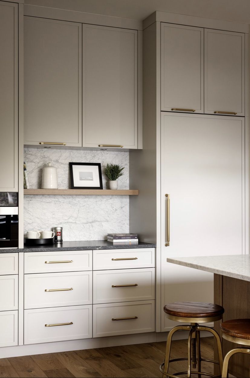 Warm ivory slim shaker cabinets with brass hardware, marble backsplash, and open wood shelf in a modern kitchen.