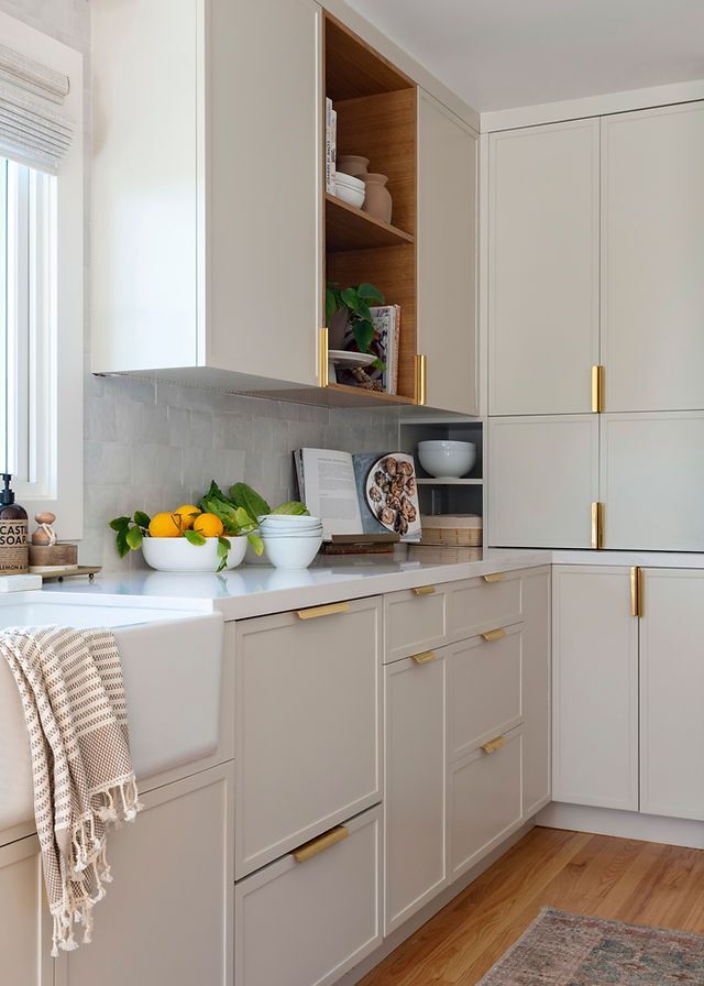 Soft white slim shaker cabinet doors with brass hardware, open wood shelving, and farmhouse sink in a bright kitchen.