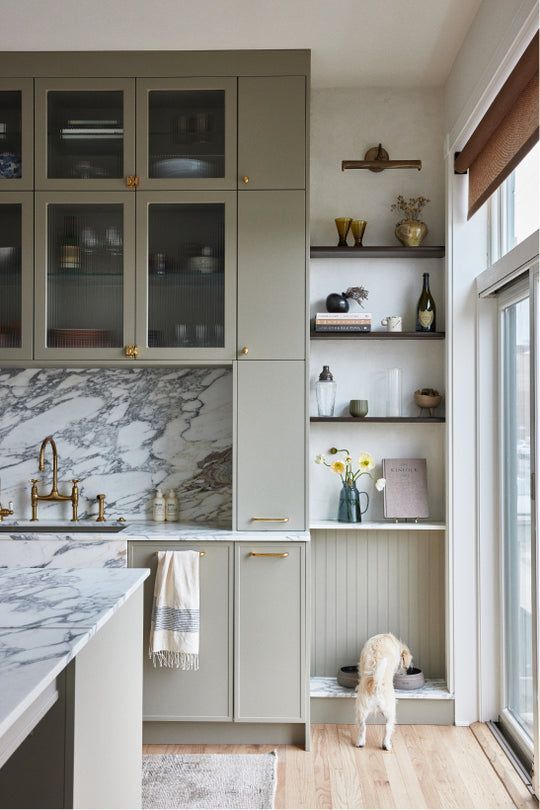 Slim shaker cabinet doors in light sage green with marble backsplash, glass upper cabinets, and brass hardware in a modern kitchen.